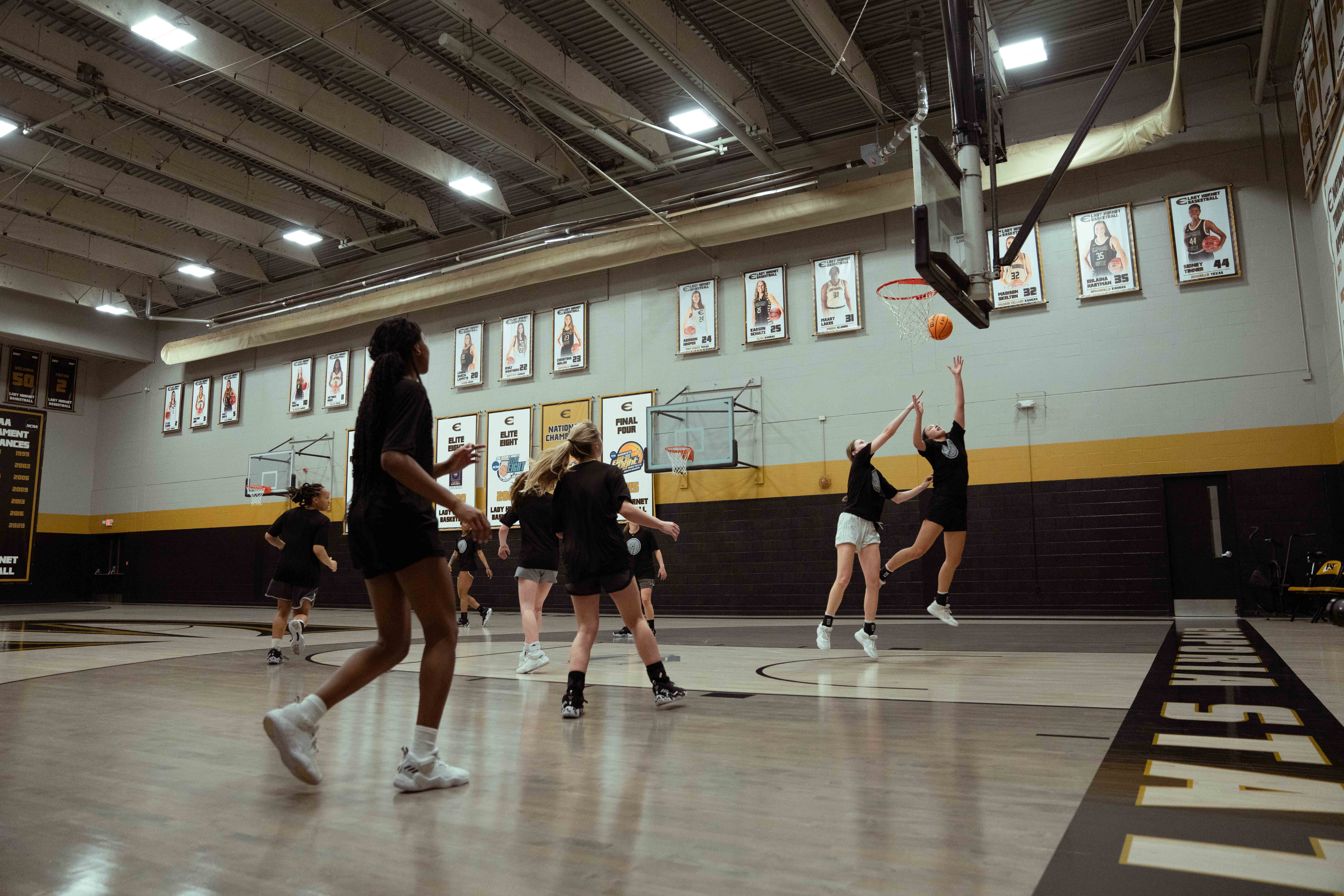 A wide shot shows a group of female basketball players practicing in a well-lit gymnasium. The players are dressed in black athletic wear, and two are actively jumping for a rebound under the hoop, one reaching towards the orange basketball. The gym features a light-colored court with "EMPORIA STATE" written along the baseline in black lettering with a yellow outline. The walls are light grey with a prominent black and yellow stripe running along the lower section. Numerous framed photos and banners, likely commemorating past athletes or achievements, adorn the walls. The ceiling has visible beams and bright fluorescent lighting.