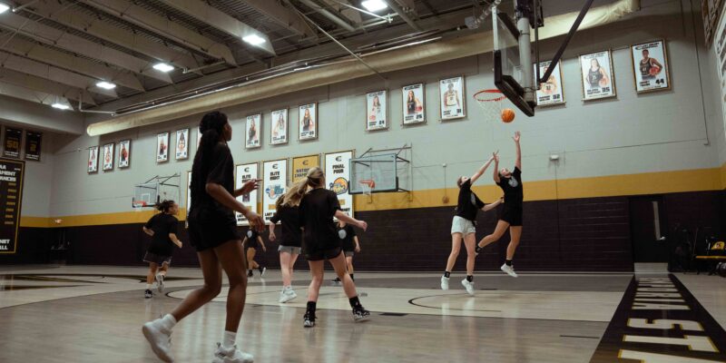 A wide shot shows a group of female basketball players practicing in a well-lit gymnasium. The players are dressed in black athletic wear, and two are actively jumping for a rebound under the hoop, one reaching towards the orange basketball. The gym features a light-colored court with "EMPORIA STATE" written along the baseline in black lettering with a yellow outline. The walls are light grey with a prominent black and yellow stripe running along the lower section. Numerous framed photos and banners, likely commemorating past athletes or achievements, adorn the walls. The ceiling has visible beams and bright fluorescent lighting.